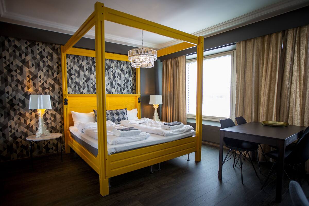 A four-poster bed with a chandelier, a dining table with chairs, and a patterned wallpaper wall.
