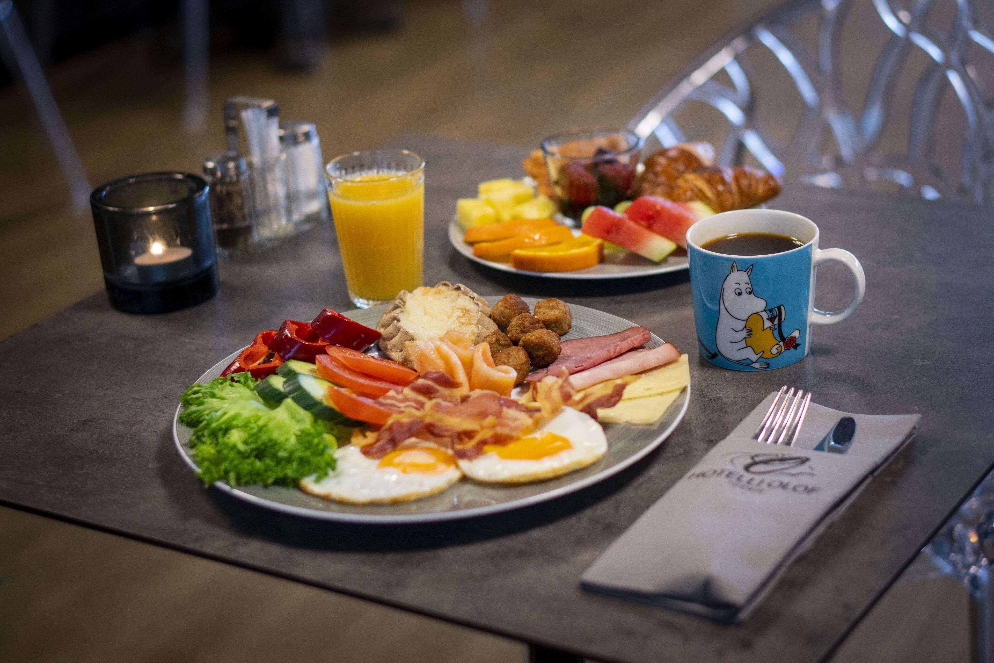 A breakfast table with plates of eggs, bacon, sausages, and toast, a glass of orange juice, and a coffee cup.