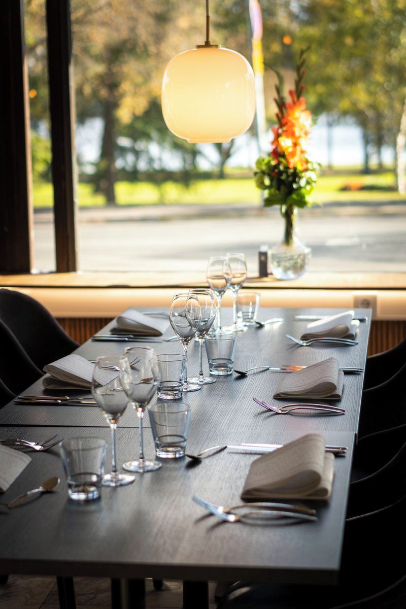 Une table à manger dressée avec des verres à vin, des couverts et des serviettes, donnant sur un jardin et agrémentée d’un luminaire suspendu.