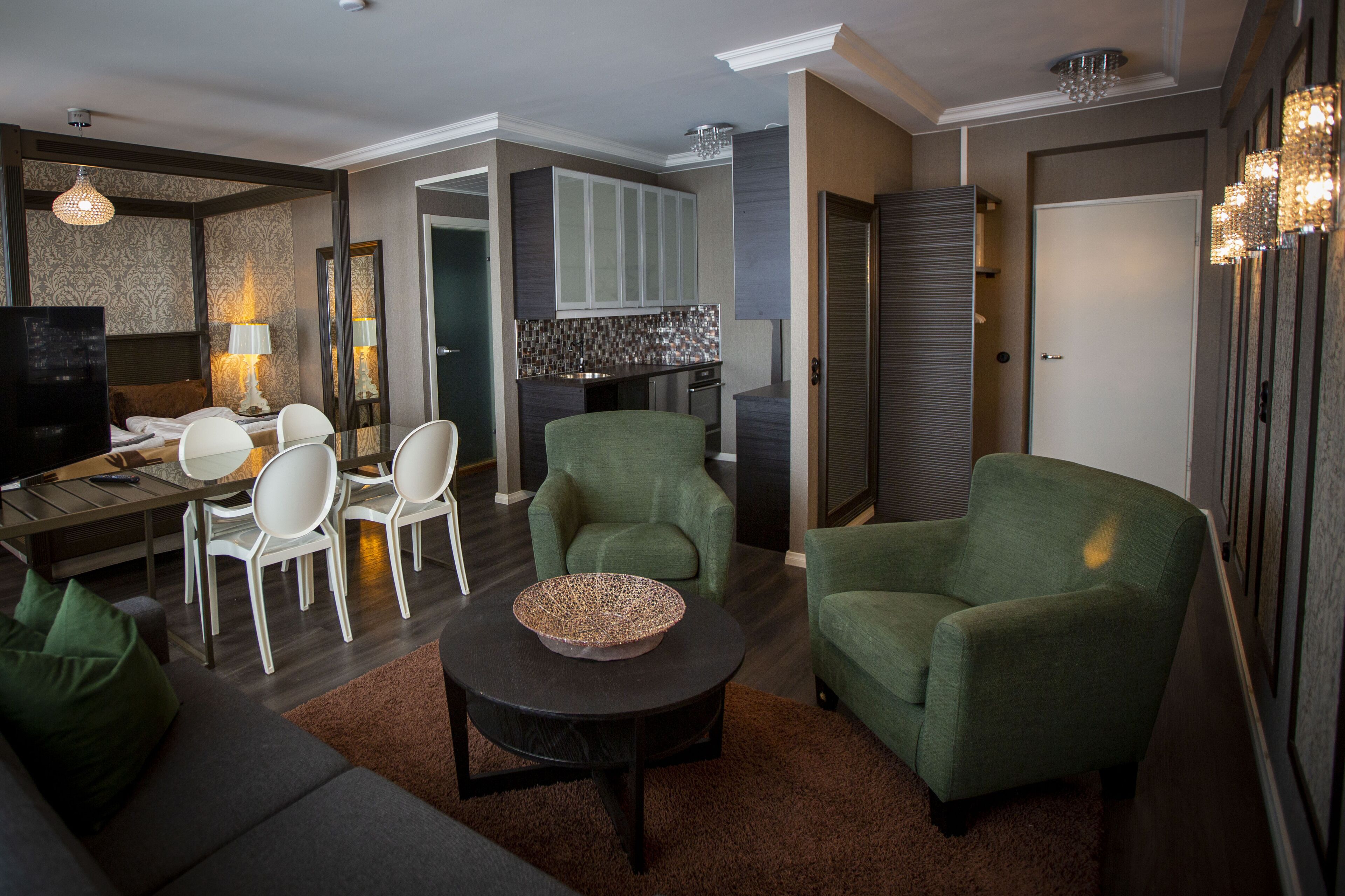 Una sala moderna con un sofá, dos sillones verdes, una mesa de comedor con sillas, un televisor y una cocina con armarios y encimera.