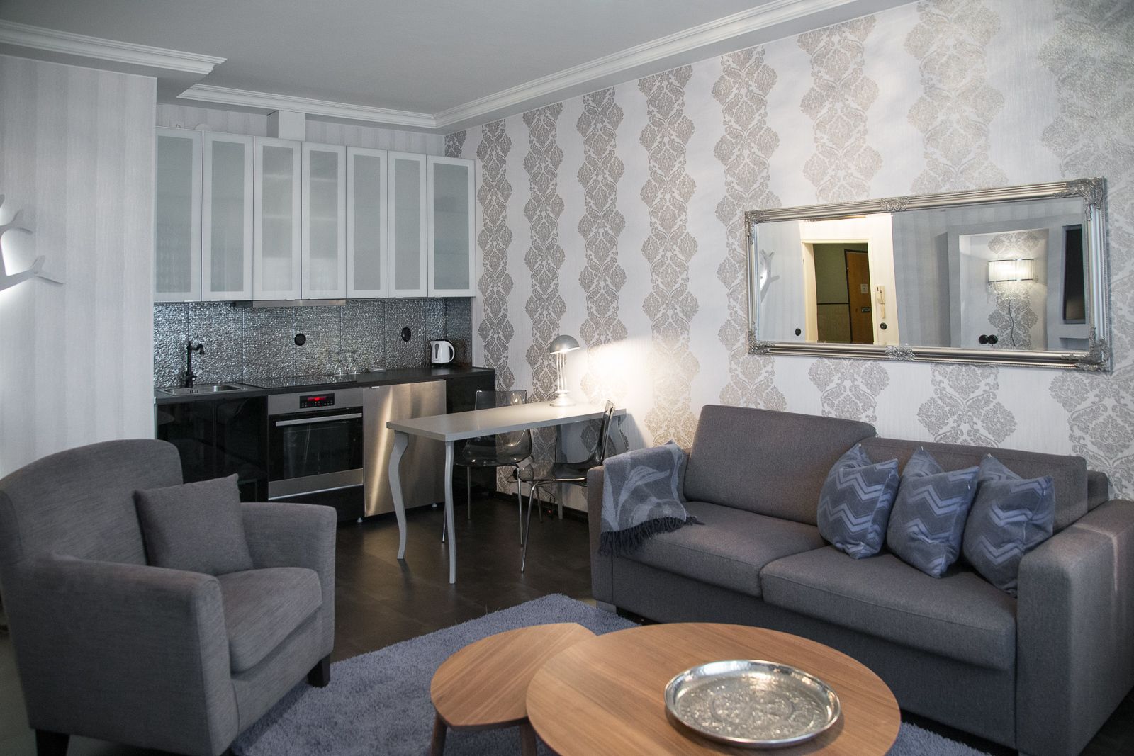 A modern living room with a grey sofa, two armchairs, a wooden coffee table, and a kitchen area with a tiled backsplash and white cabinets.