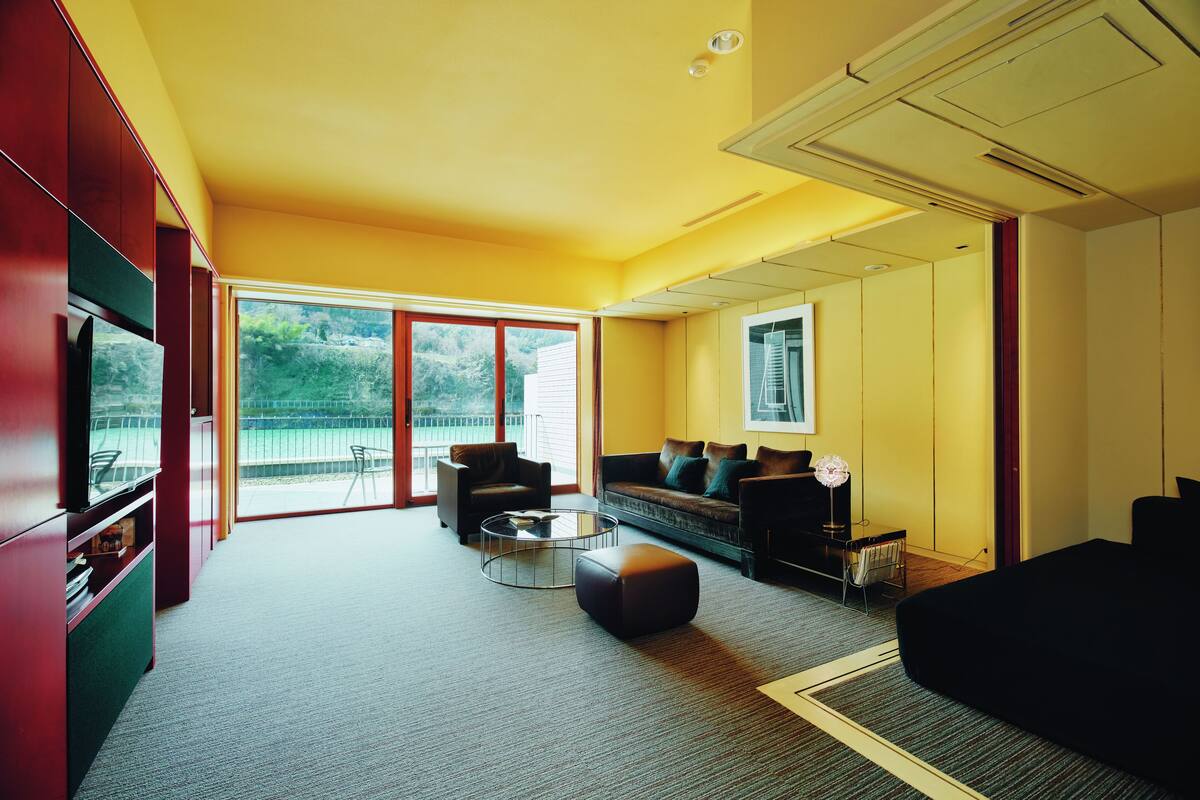 A modern living room with a sofa, a coffee table, and a TV. There is a balcony with a view of greenery.