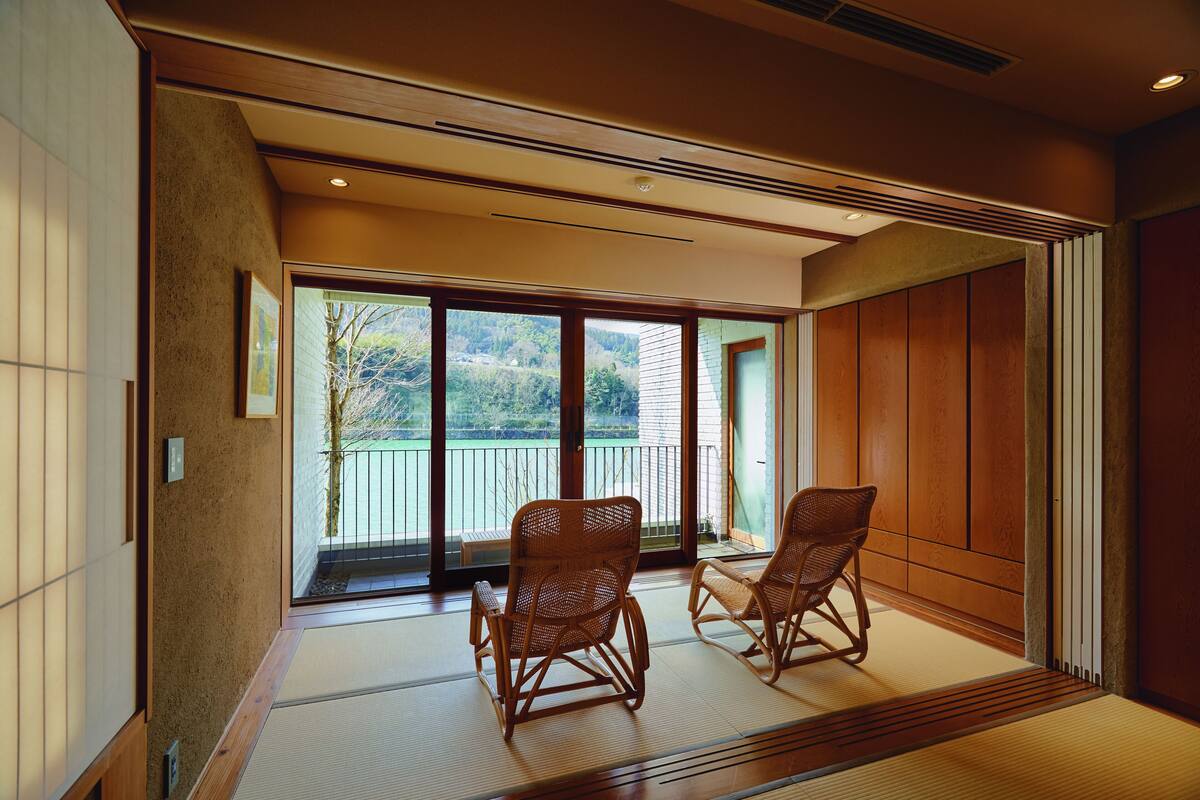 A traditional Japanese room with tatami flooring, two wicker chairs, a sliding glass door leading to a balcony, and wooden paneling.
