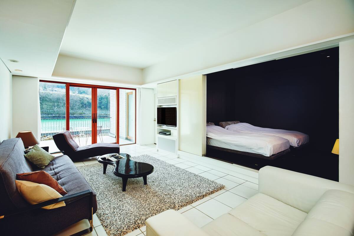 A modern hotel room with a large bed, a sofa, a coffee table, and a TV. There is a balcony with a view of the water.