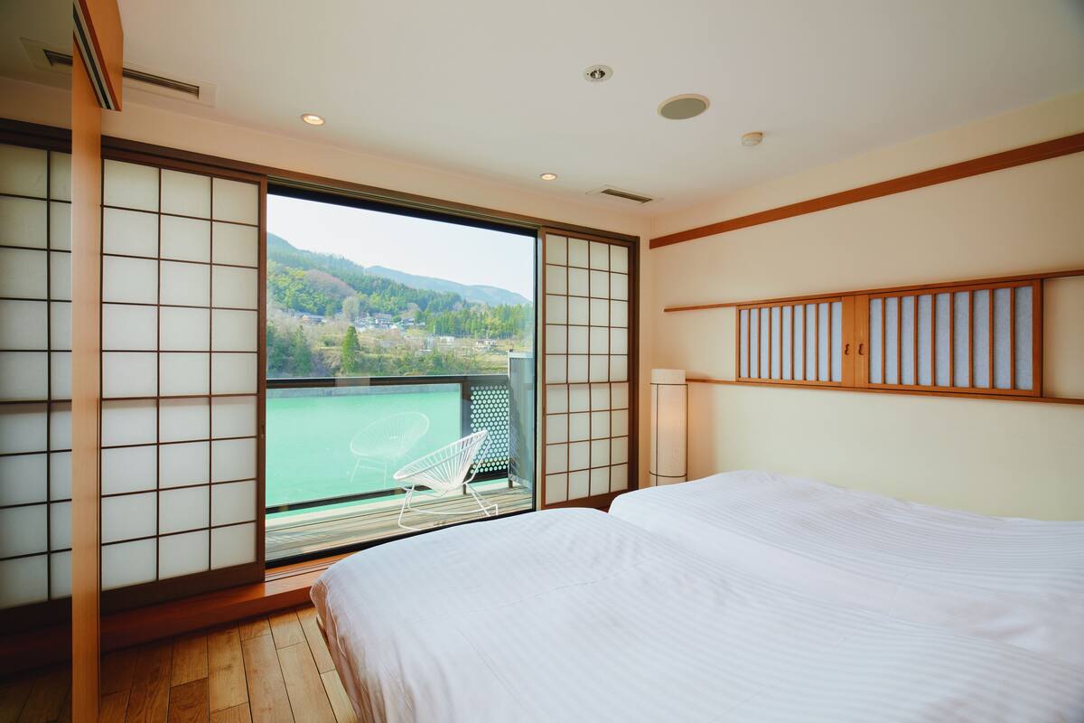 A bedroom with a large window, sliding shoji doors, a bed, and a view of a tennis court and mountains.