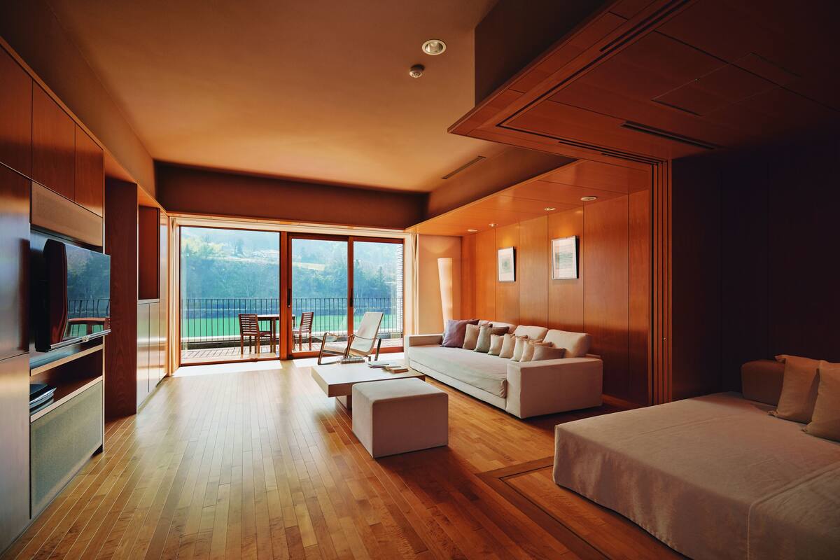 A modern living room with a large sofa, a coffee table, and a TV. There is a balcony with a view of the outdoors.