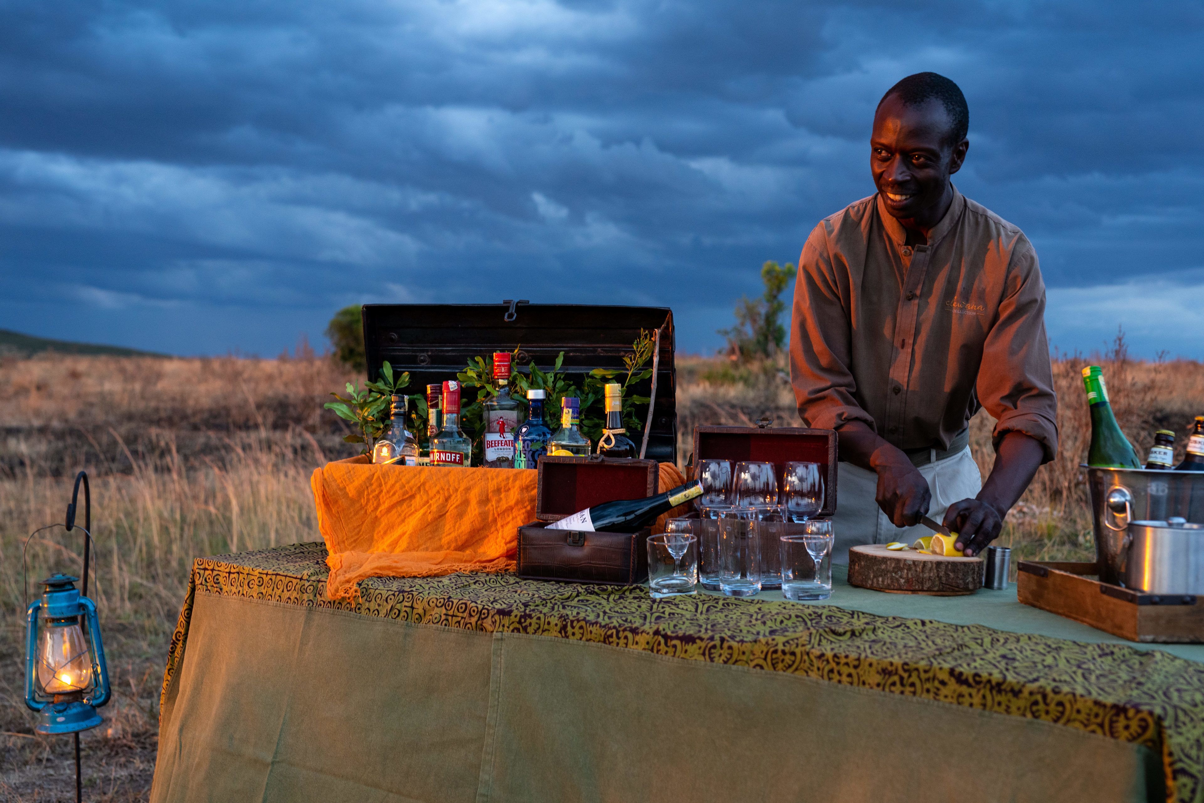 A table set outdoors with a variety of bottles and glasses, a lantern, and a person preparing food.