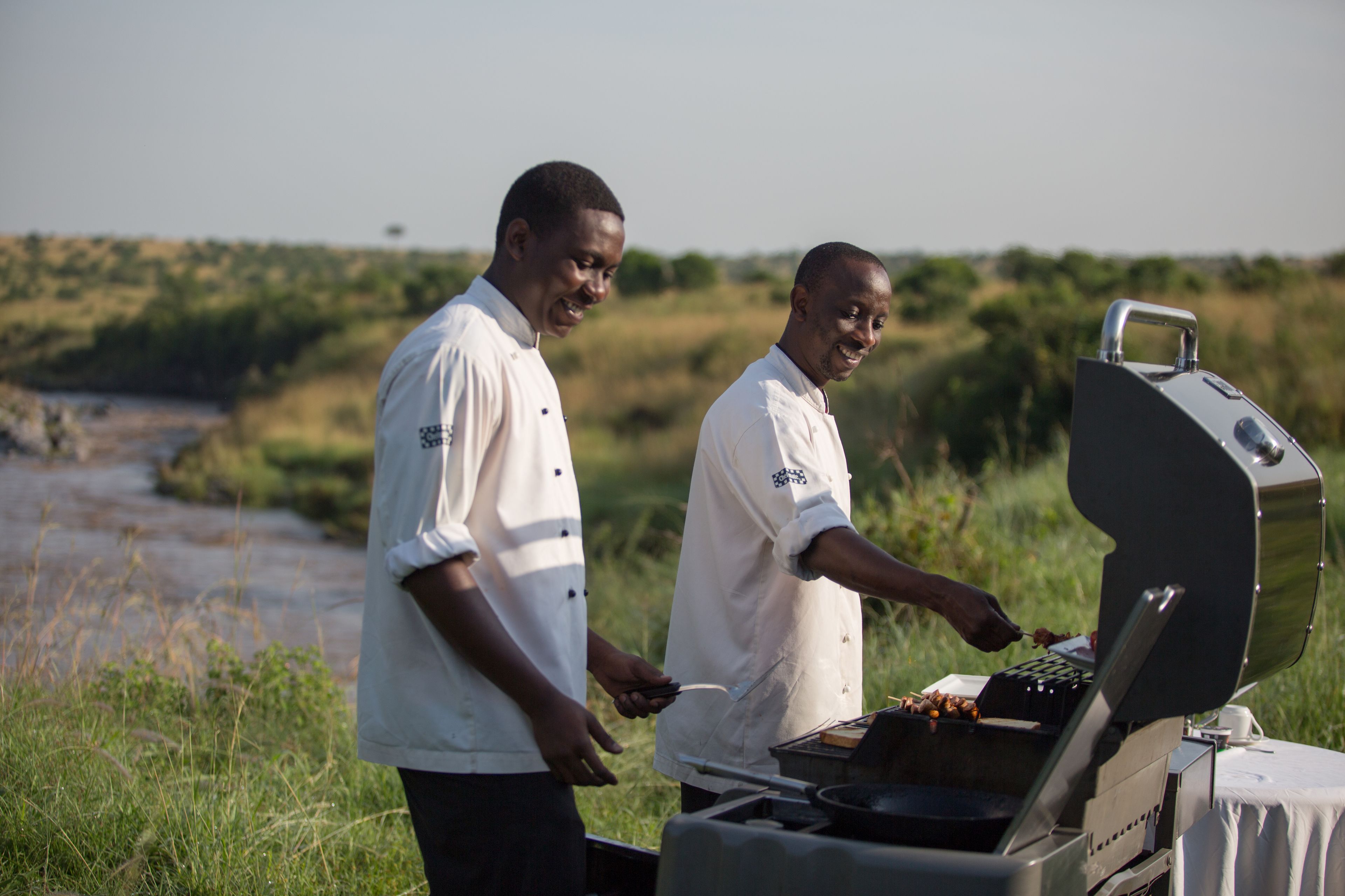Two men grilling outdoors with a river in the background.