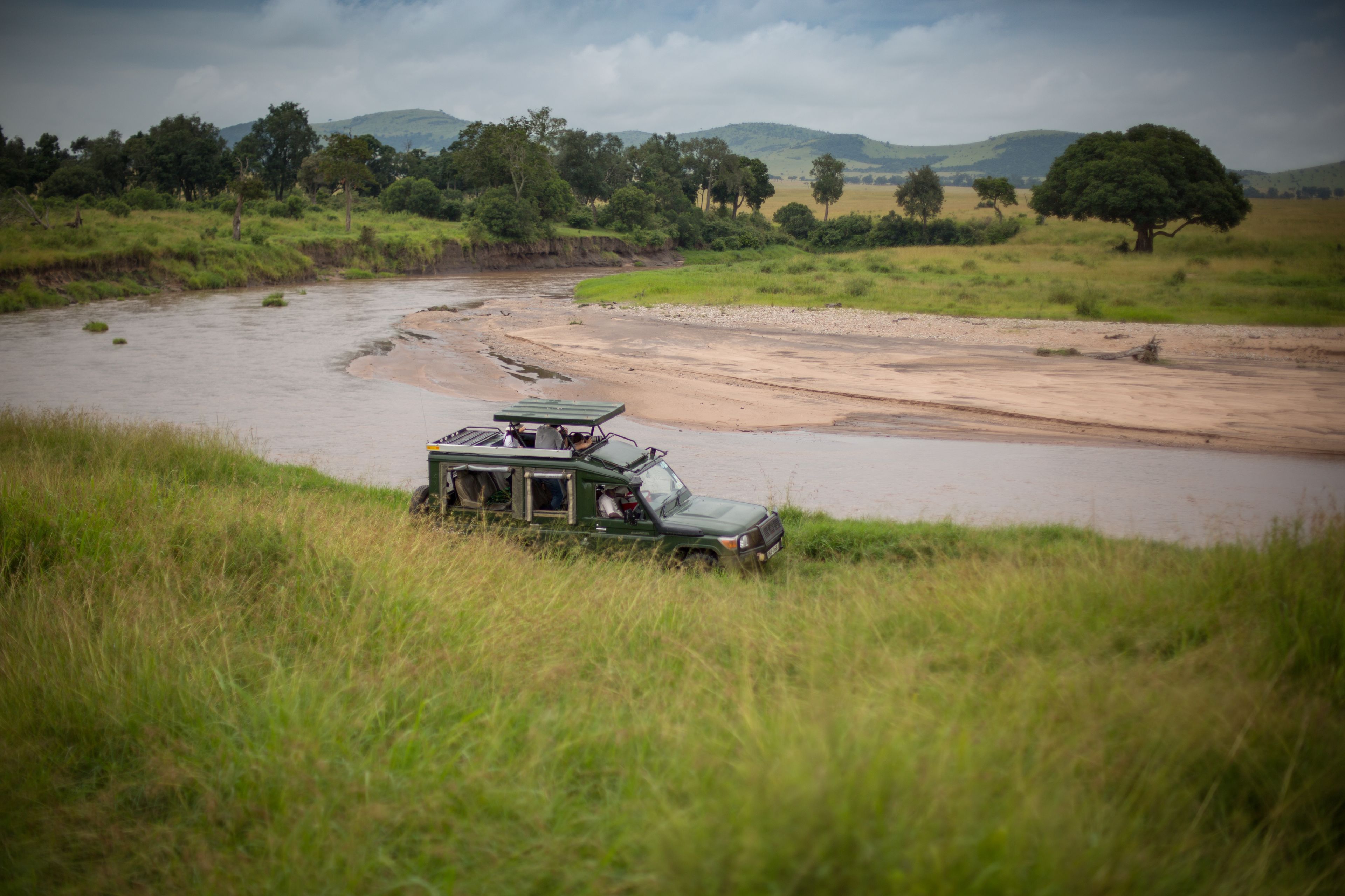 Un véhicule de safari vert est garé au bord d’une rivière, avec un paysage verdoyant et des arbres en arrière-plan.