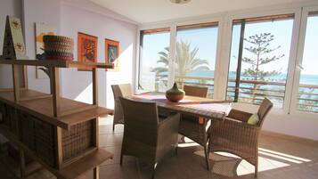 A dining area with wicker furniture, a table, and a view of the sea through large windows.