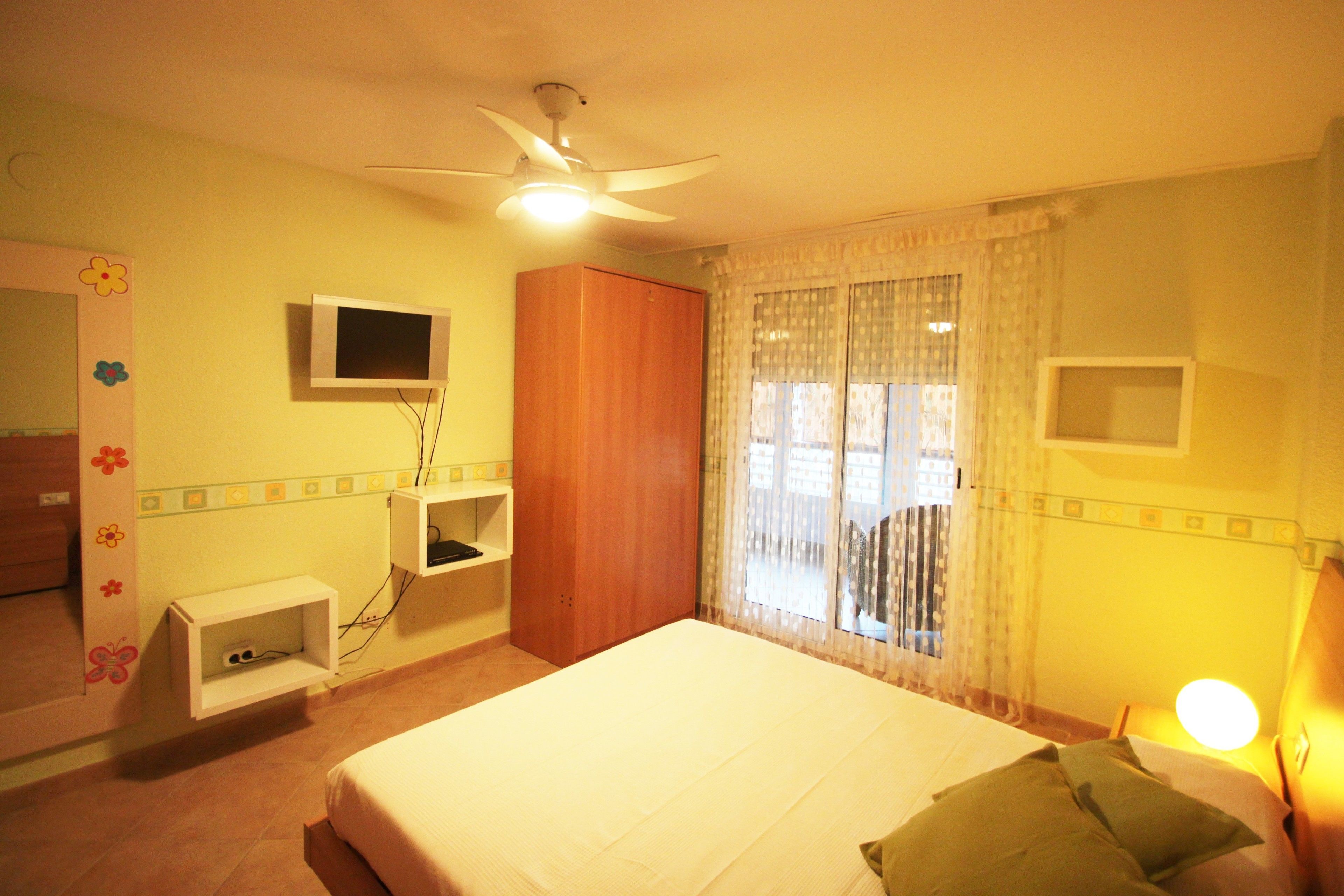 A bedroom with a bed, a ceiling fan, a TV, a wardrobe, and a window with curtains.