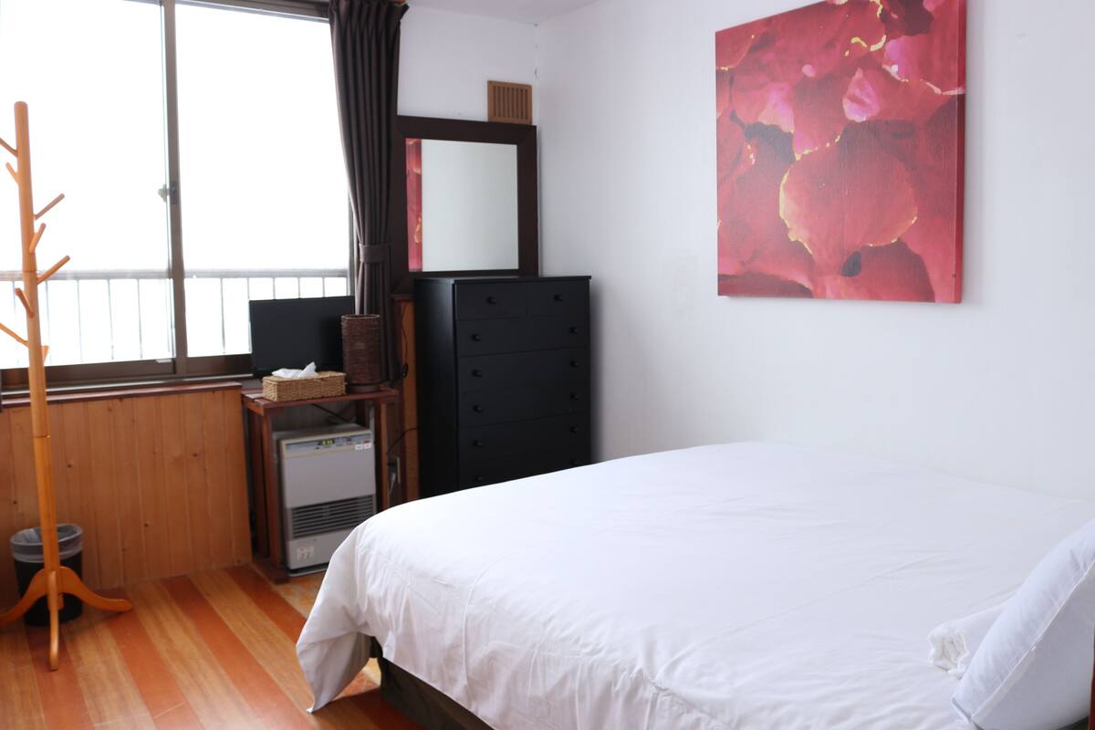 Une chambre à coucher avec un lit, une commode, un radiateur, une fenêtre avec des rideaux et un tableau accroché au mur.