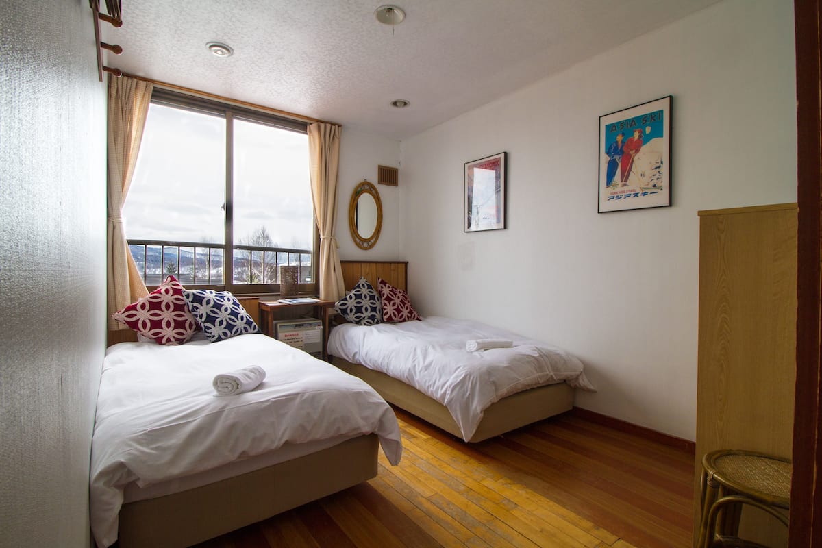 Une chambre d’hôtel avec deux lits, une fenêtre avec des rideaux, un plancher en bois, un miroir et un tableau encadré au mur.