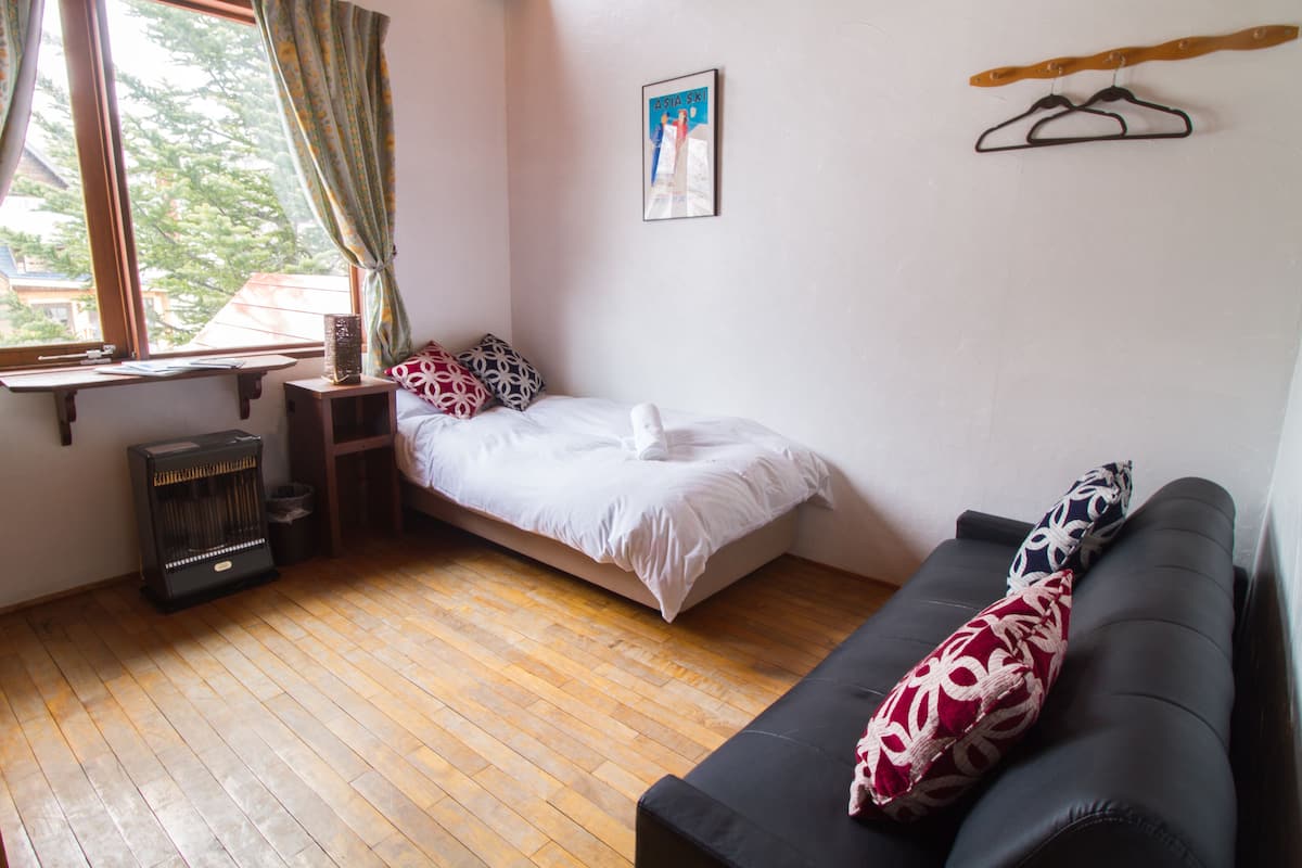 Une pièce avec un lit, un canapé noir, un plancher en bois, une fenêtre avec des rideaux, un tableau au mur et un radiateur.
