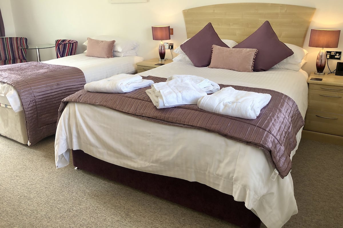 A hotel room with two beds, each with a headboard, pillows, and bedside lamps. There are towels on the beds and a carpeted floor.