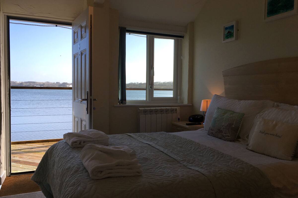 A bedroom with a bed, a window with a view of the sea, a radiator, and a bedside table with a lamp.