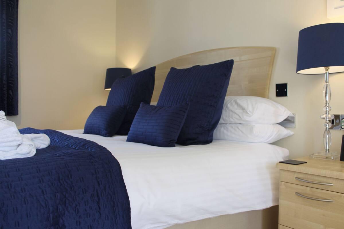 A neatly made bed with a headboard, a bedside lamp, and a small bedside table with a phone and a box.