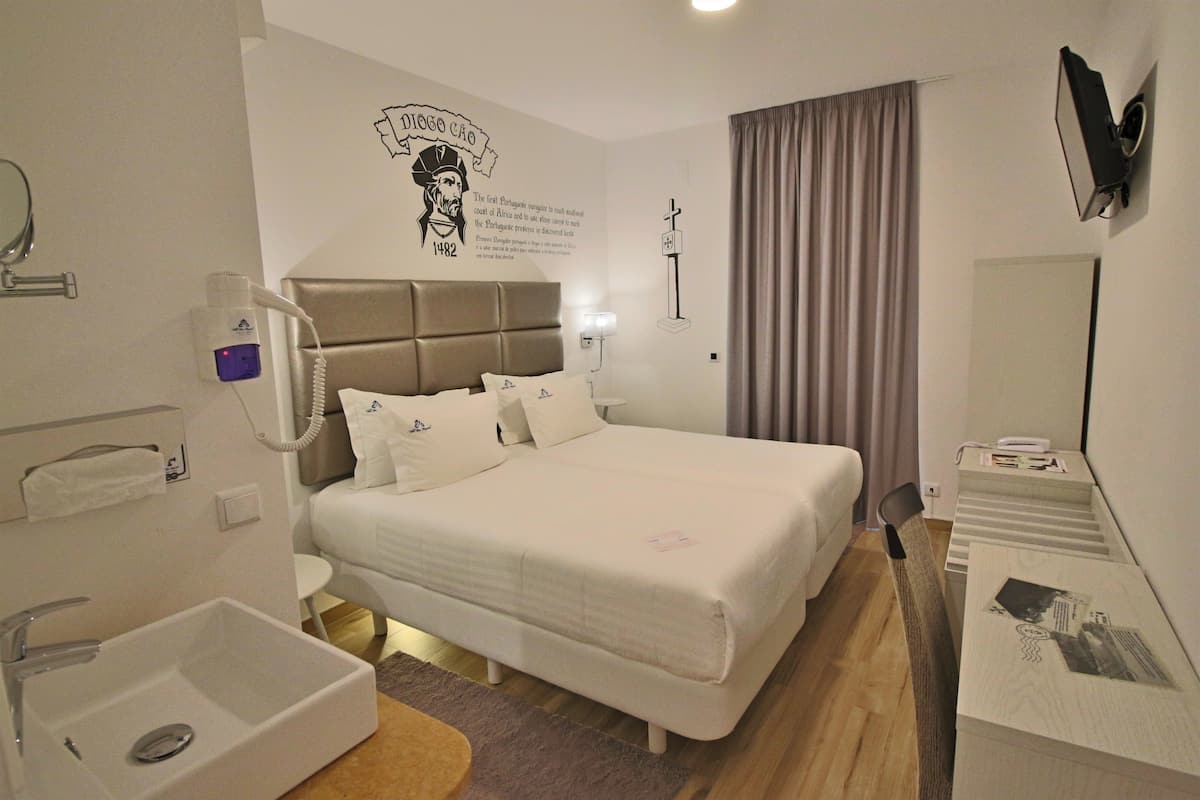 Une chambre d’hôtel comprenant un lit, une table de chevet, un miroir, un téléviseur fixé au mur, une fenêtre avec des rideaux et un élément décoratif mural.