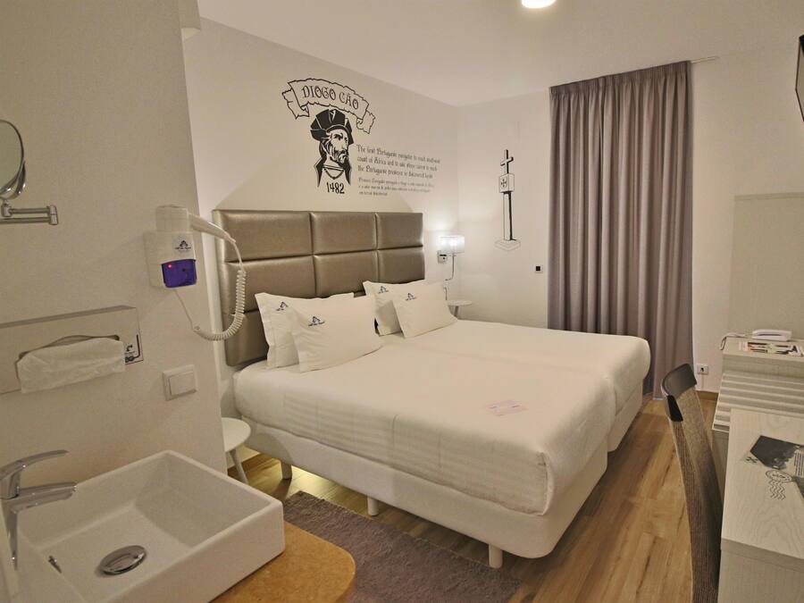 Habitación de hotel con cama, mesita de noche, espejo, televisor montado en la pared, ventana con cortinas y un adorno en la pared.