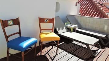 Una terraza de madera con dos sillas, una con cojín azul y otra con cojín amarillo, y una pequeña mesa con una planta.