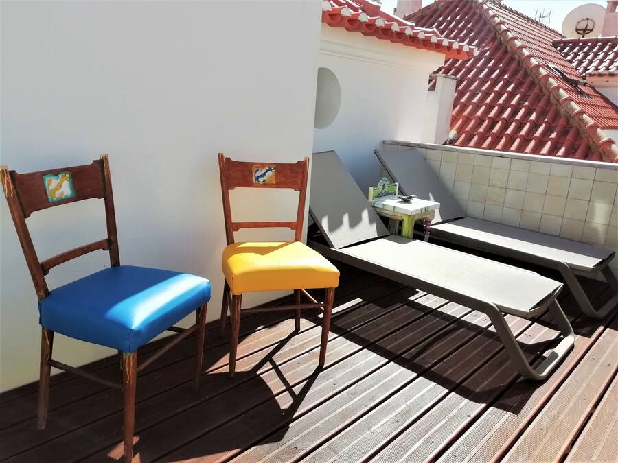 Una terraza de madera con dos sillas, una con cojín azul y otra con cojín amarillo, y una pequeña mesa con una planta.