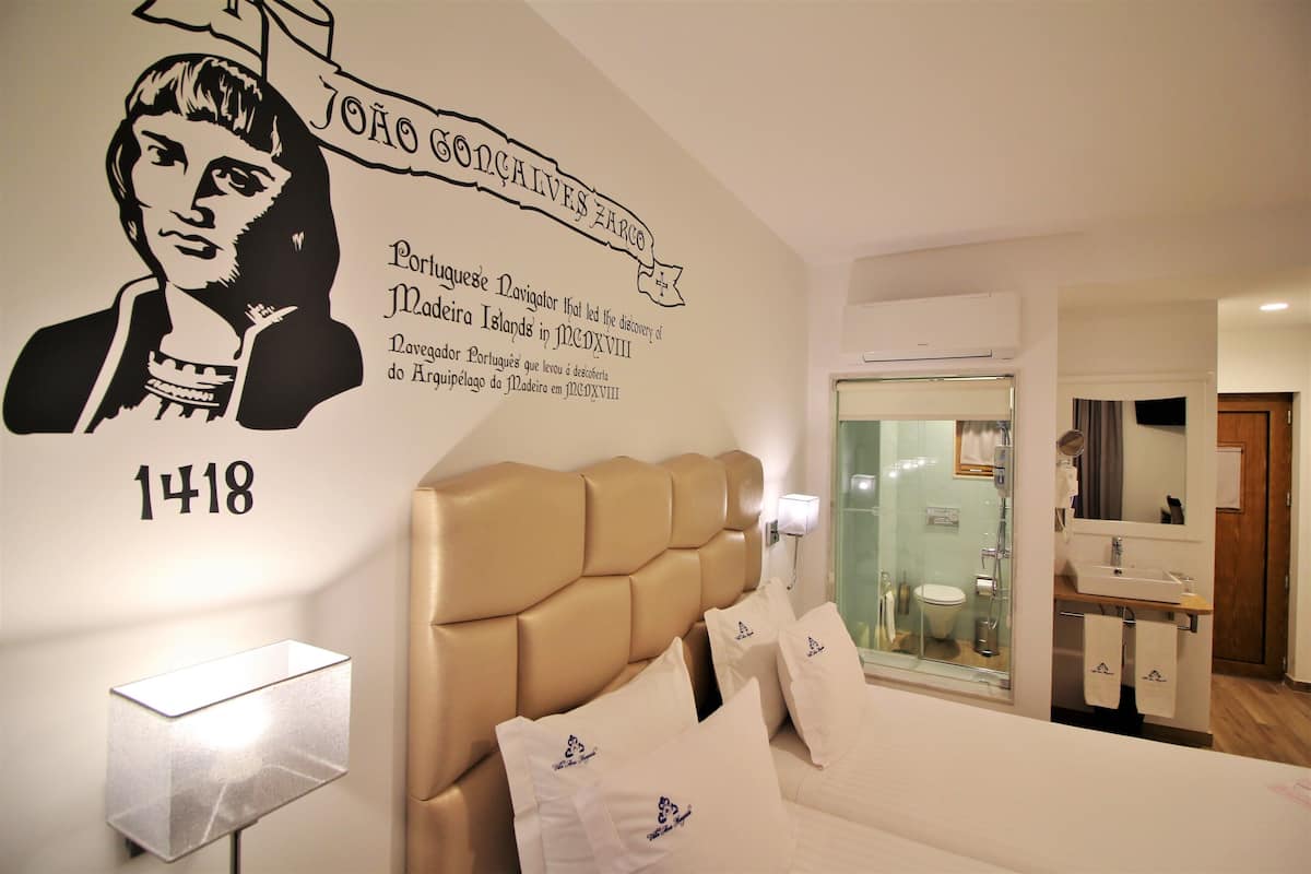 Une chambre d’hôtel avec un grand lit, une tête de lit, une lampe fixée au mur et une salle de bain visible à travers une porte en verre.
