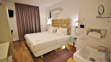 Una habitación de hotel con cama, mesita de noche, lámpara, espejo, lavabo y dispensador de toallas en la pared.