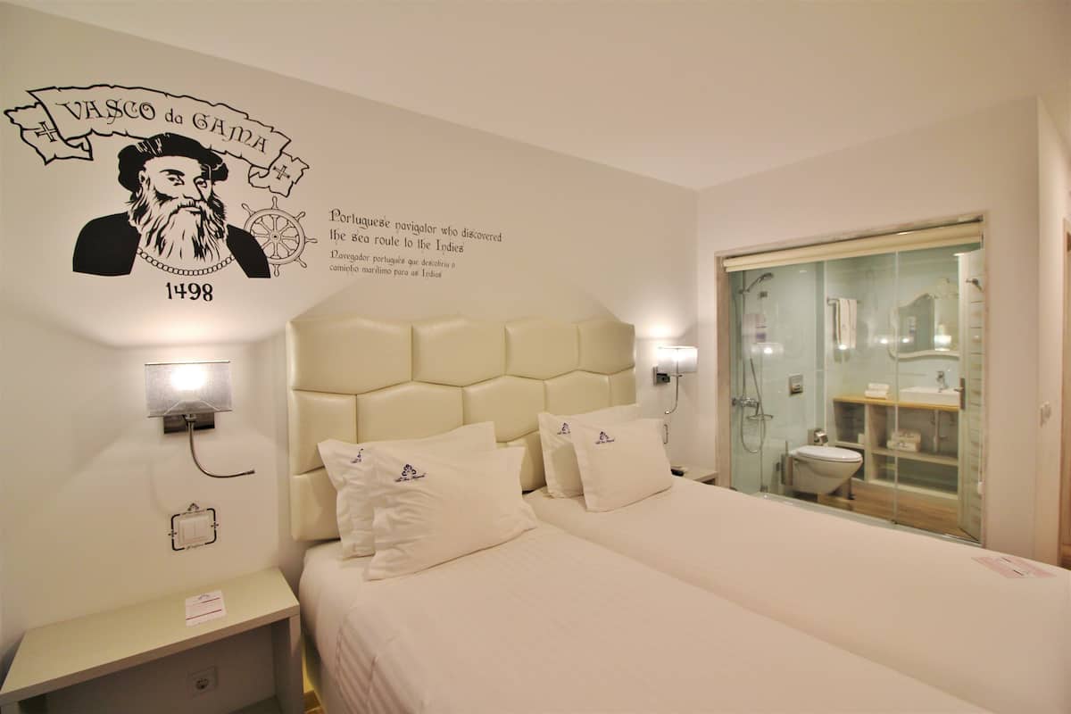Une chambre d’hôtel avec un grand lit, une tête de lit, une table de chevet, un élément de décoration murale et une salle de bain visible à travers une porte en verre.