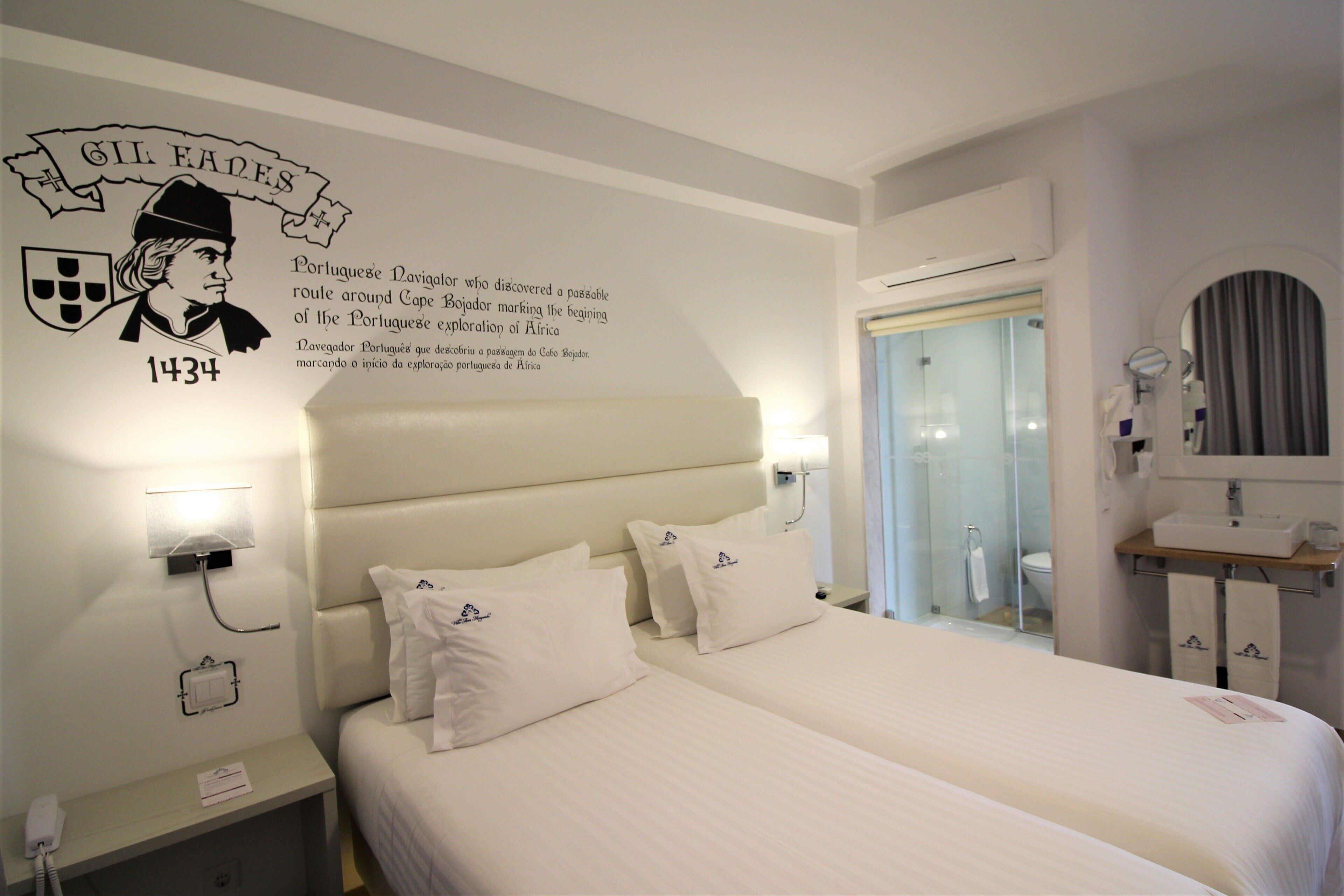 Une chambre d’hôtel avec deux lits, une table de chevet, un téléphone fixé au mur et une salle de bain visible à travers une porte en verre.