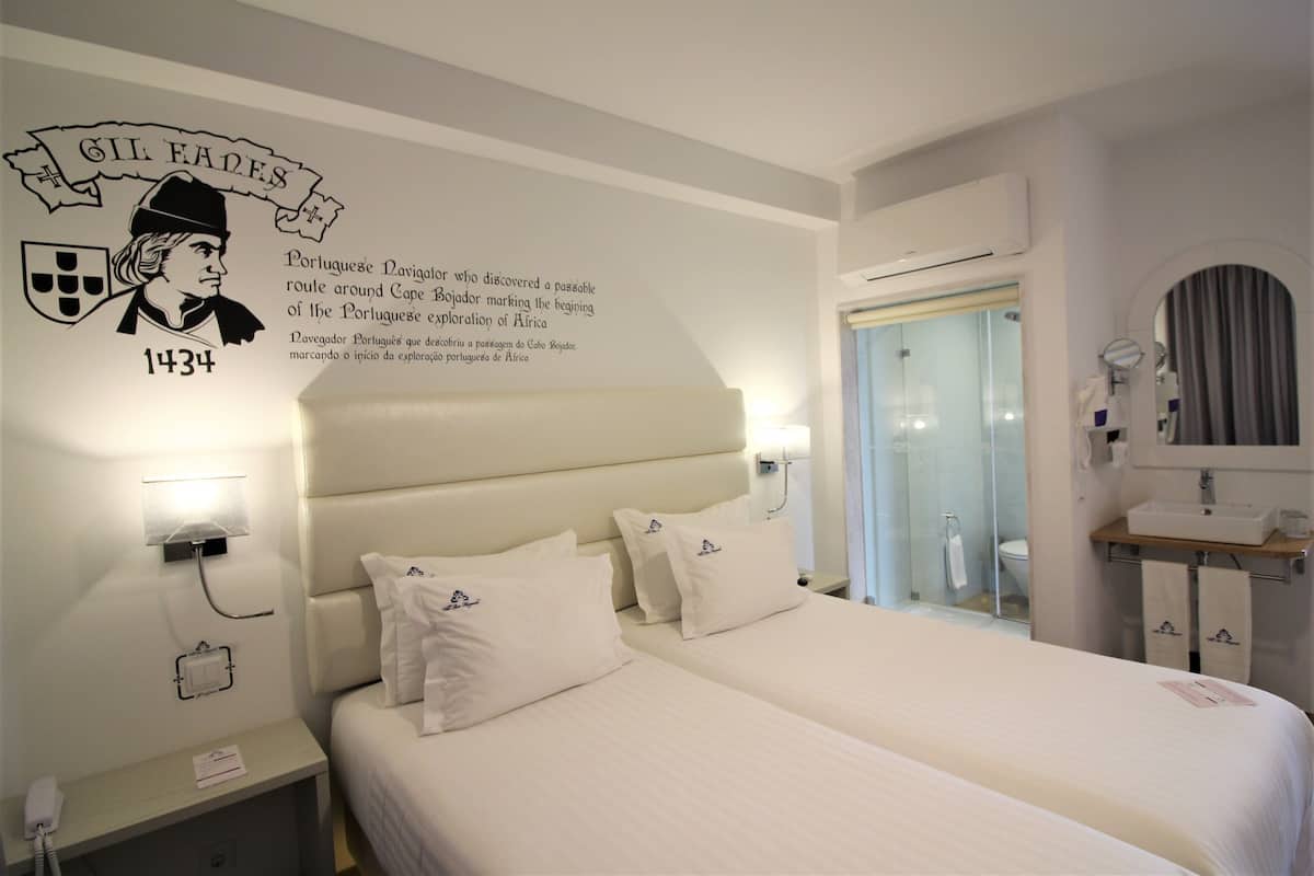 Une chambre d’hôtel avec deux lits, une table de chevet, un téléphone fixé au mur et une salle de bain visible à travers une porte en verre.