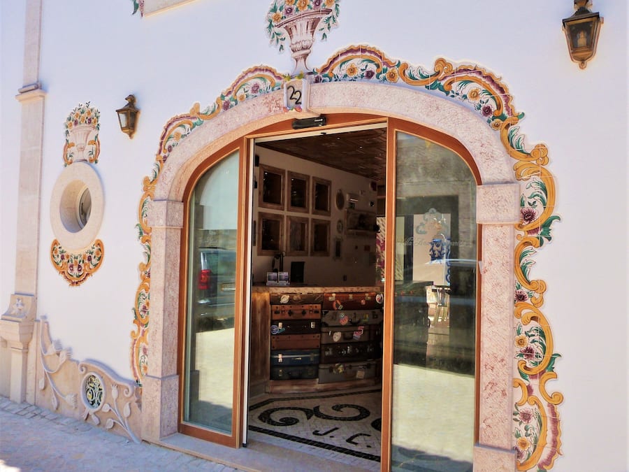 Entrada de un hotel boutique con diseños florales y geométricos ornamentales, un amplio arco de entrada y una exhibición de maletas antiguas en el interior.