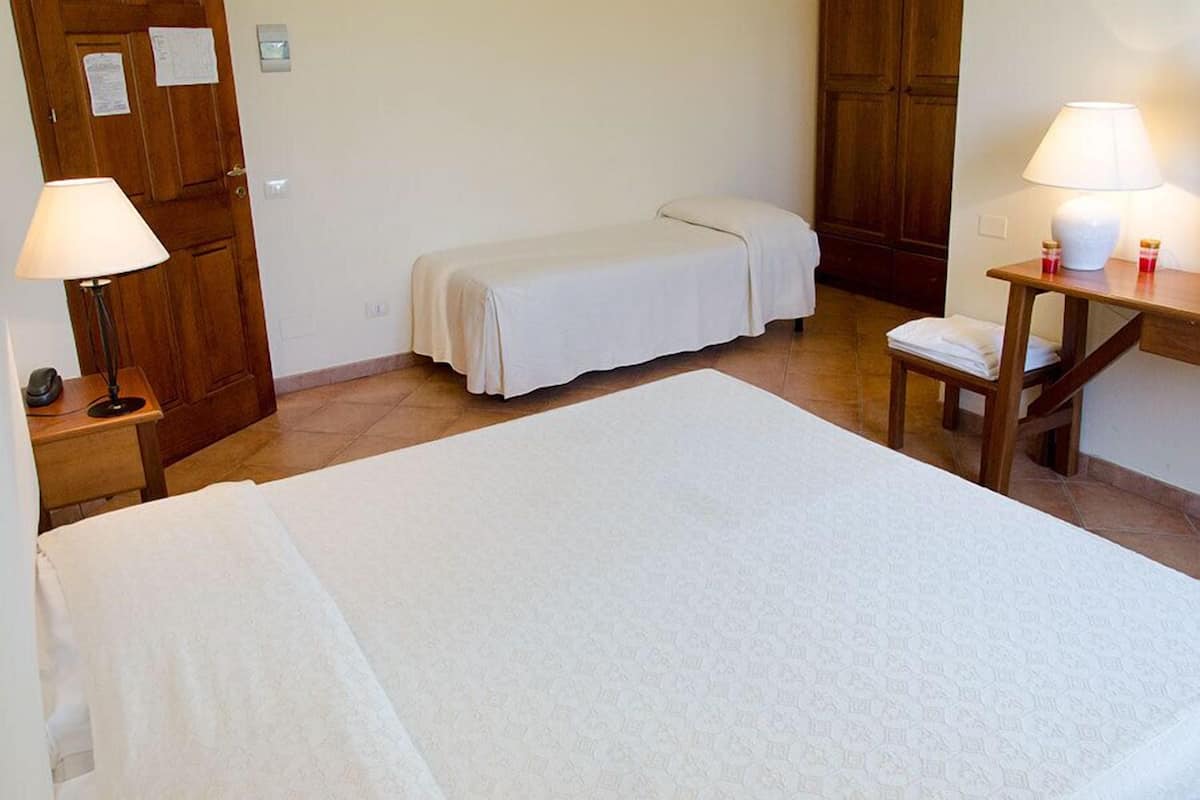 A hotel room with a large bed, a bedside table with a lamp, a small bench, and a wooden wardrobe.