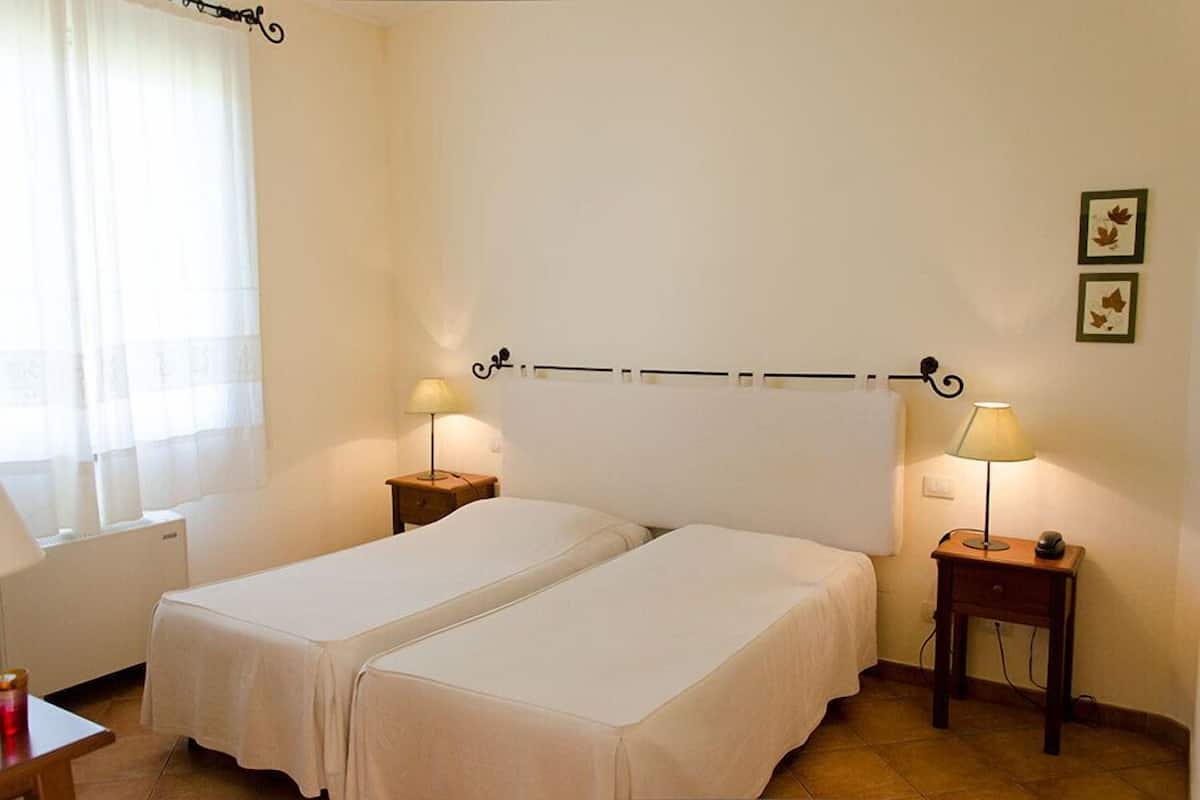 A room with two single beds, a window with sheer curtains, a radiator, and two bedside tables with lamps.