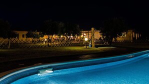 A swimming pool at night with a fence and a building in the background. - Hotel Tanca Irde (Capoterra)
