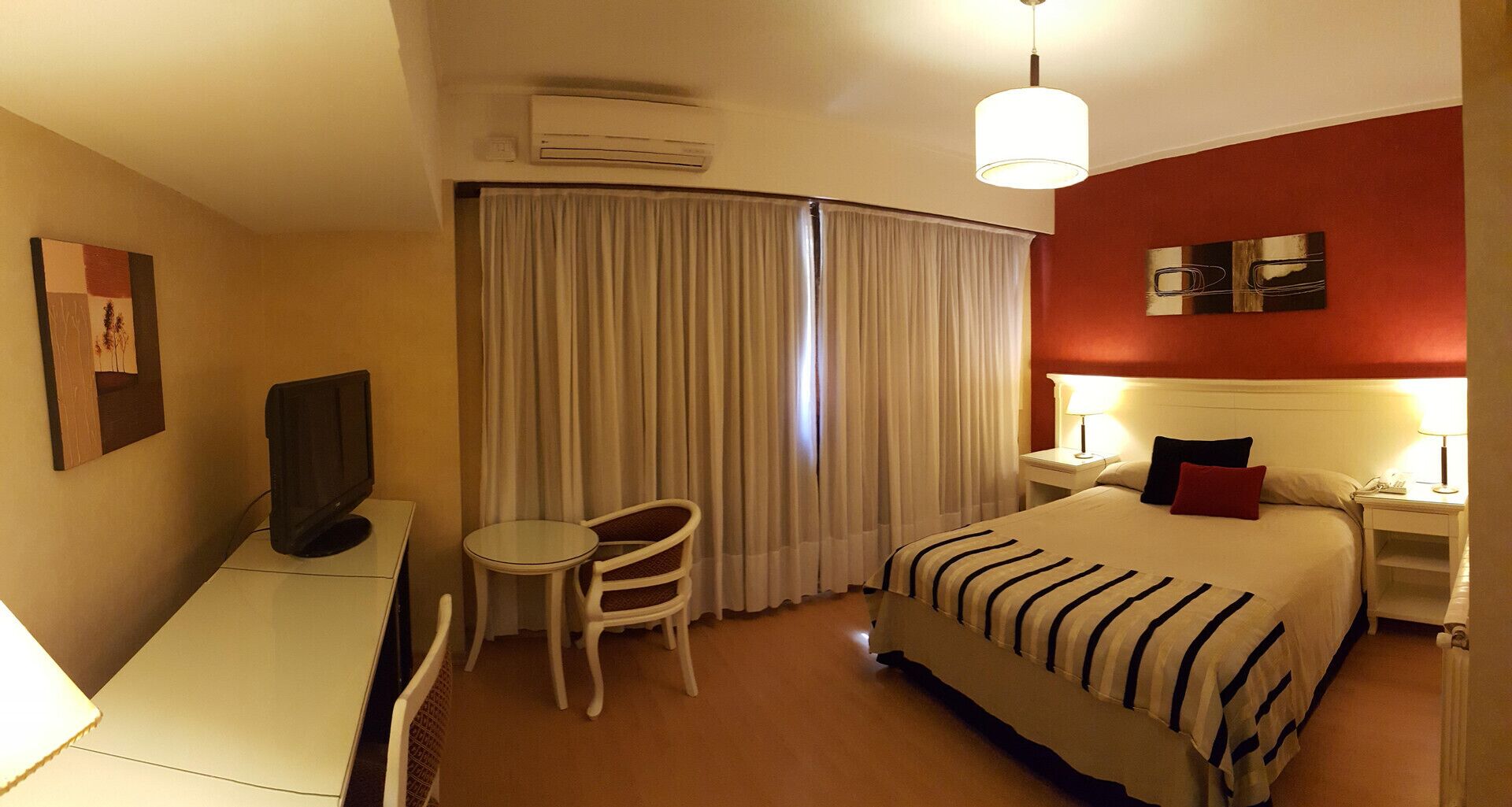 A hotel room with a bed, a desk with a TV, a chair, a nightstand with a lamp, and a window with curtains.