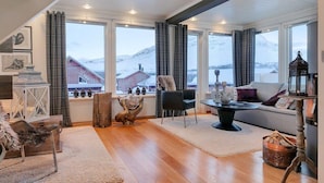 A cozy living room with a wooden floor, a sofa, a coffee table, and a view of snow-covered mountains through large windows. - RIBO Apartment Katterjokk (Riksgränsen)