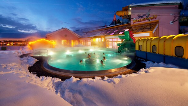 Ein beheizter Außenpool, umgeben von Schnee, mit einem Gebäude im Hintergrund, auf dem 'Wasservelt' steht.