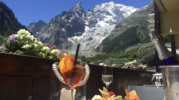 A table with a fruit platter and drinks, a bottle of champagne, and a view of snow-capped mountains. - Shatush Hotel (Courmayeur)