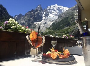 A table with a fruit platter and drinks, a bottle of champagne, and a view of snow-capped mountains. - Shatush Hotel (Courmayeur)