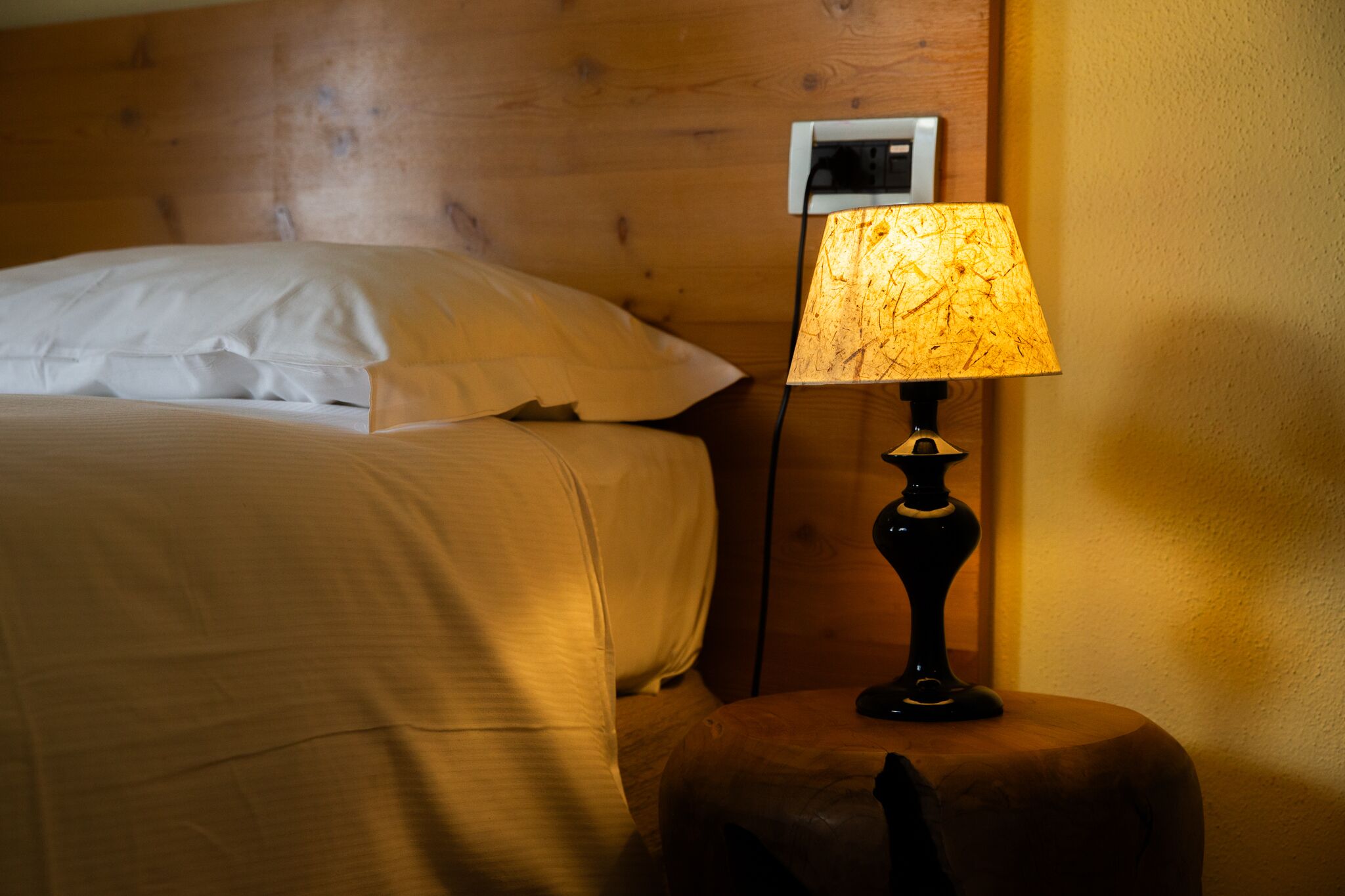 Una cama bien tendida con sábanas blancas, cabecera de madera y mesita de noche con lámpara.