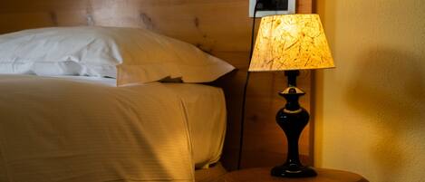 Una cama bien tendida con sábanas blancas, cabecera de madera y mesita de noche con lámpara.
