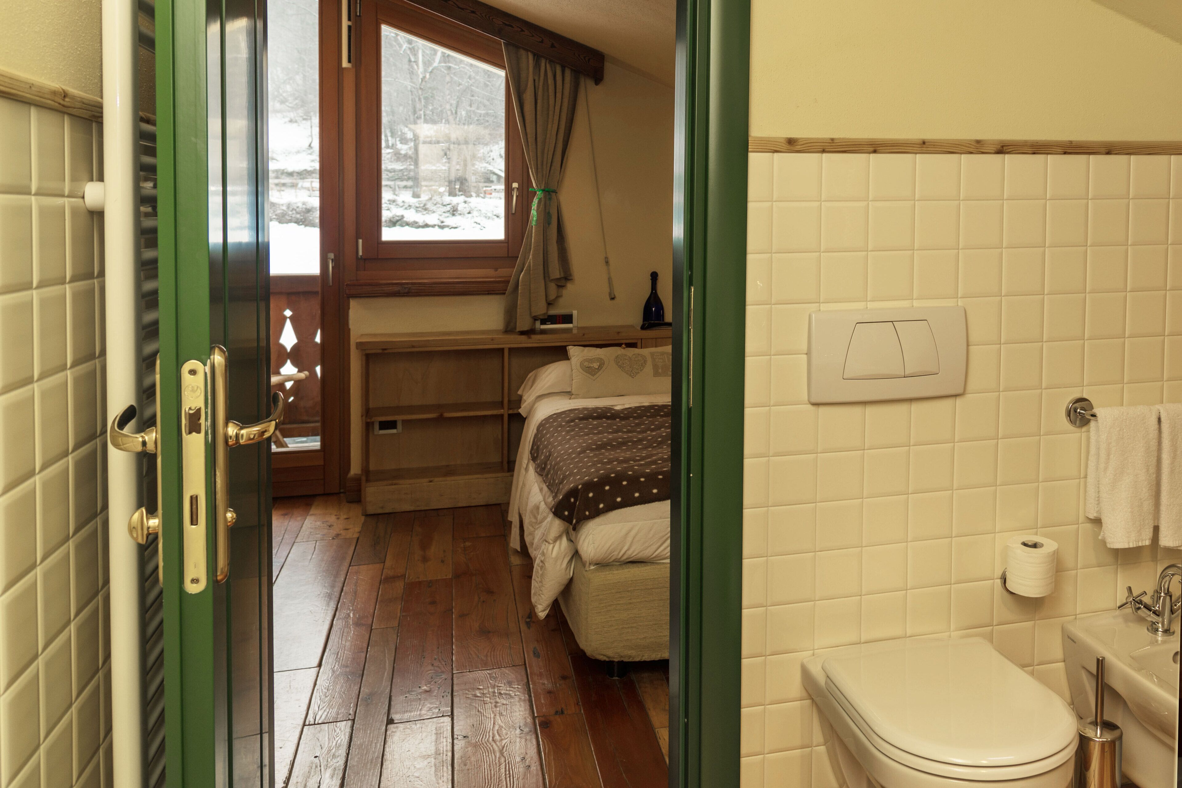 A room with a wooden floor, a bed with a brown and white polka dot blanket, a window with curtains, a wooden shelf with a bottle, and a white toilet with a paper towel holder.