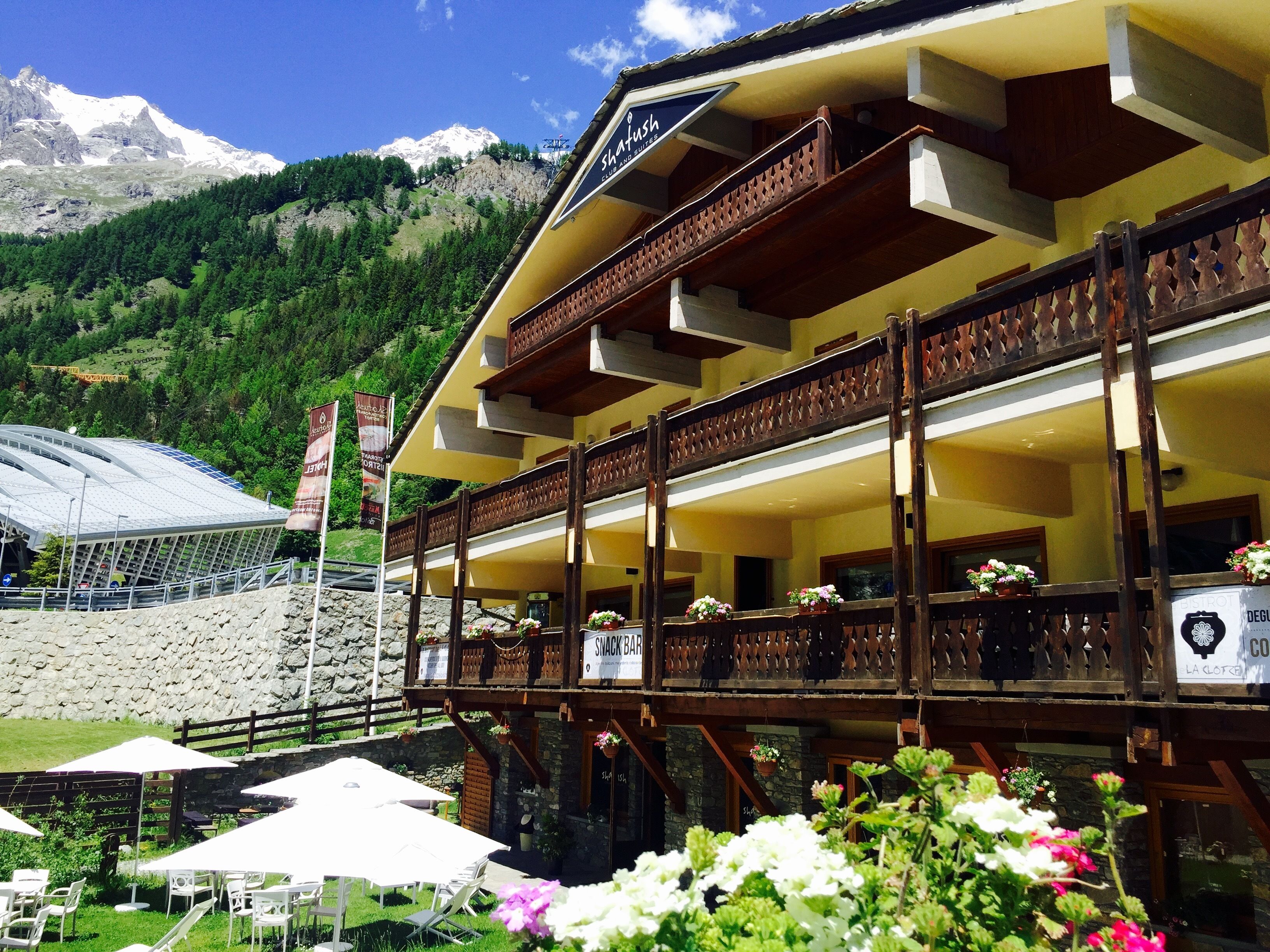 A chalet-style building with a balcony, outdoor seating area, and a mountain backdrop.