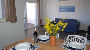 A dining area with a table set for two, a vase with yellow flowers, a blue sofa, and a painting of a landscape on the wall. - Gorgonia Suites (Sali)