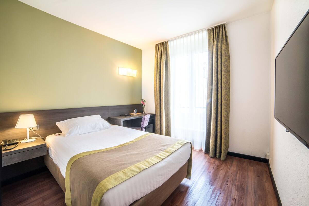 A hotel room with a bed, bedside table, lamp, and a large window with curtains.