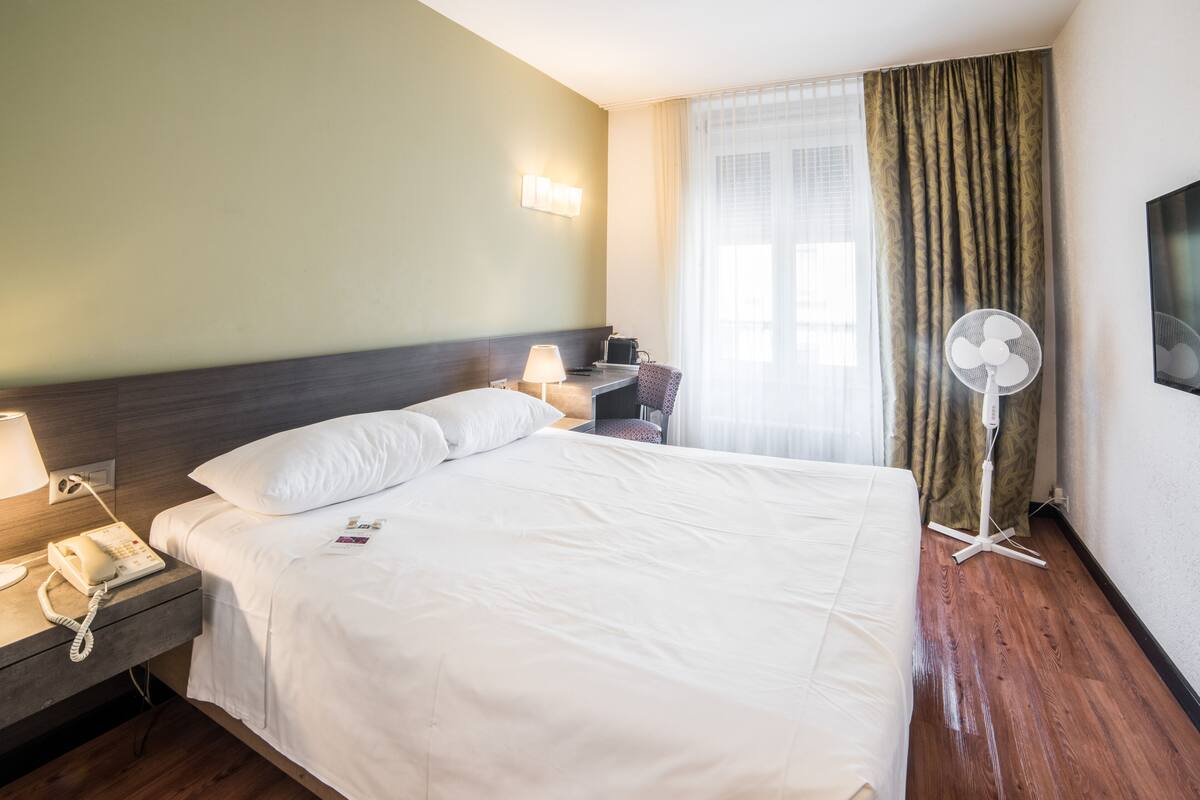A hotel room with a large bed, bedside table with a phone, lamp, and a small table with a chair. There is a standing fan and a window with curtains.