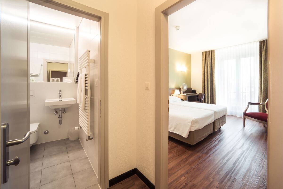A hotel room with a bed, a bedside table, a chair, a window with curtains, a bathroom with a sink and toilet, and a mirror.