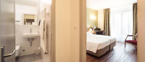 A hotel room with a bed, a bedside table, a chair, a window with curtains, a bathroom with a sink and toilet, and a mirror.