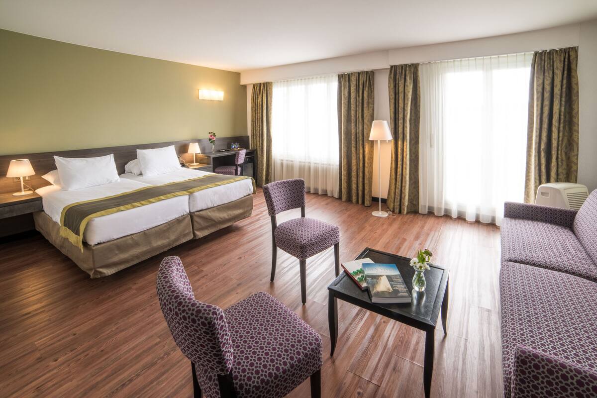 A hotel room with a double bed, two chairs, a small table with a book and flowers, a desk with a lamp, and a sofa.