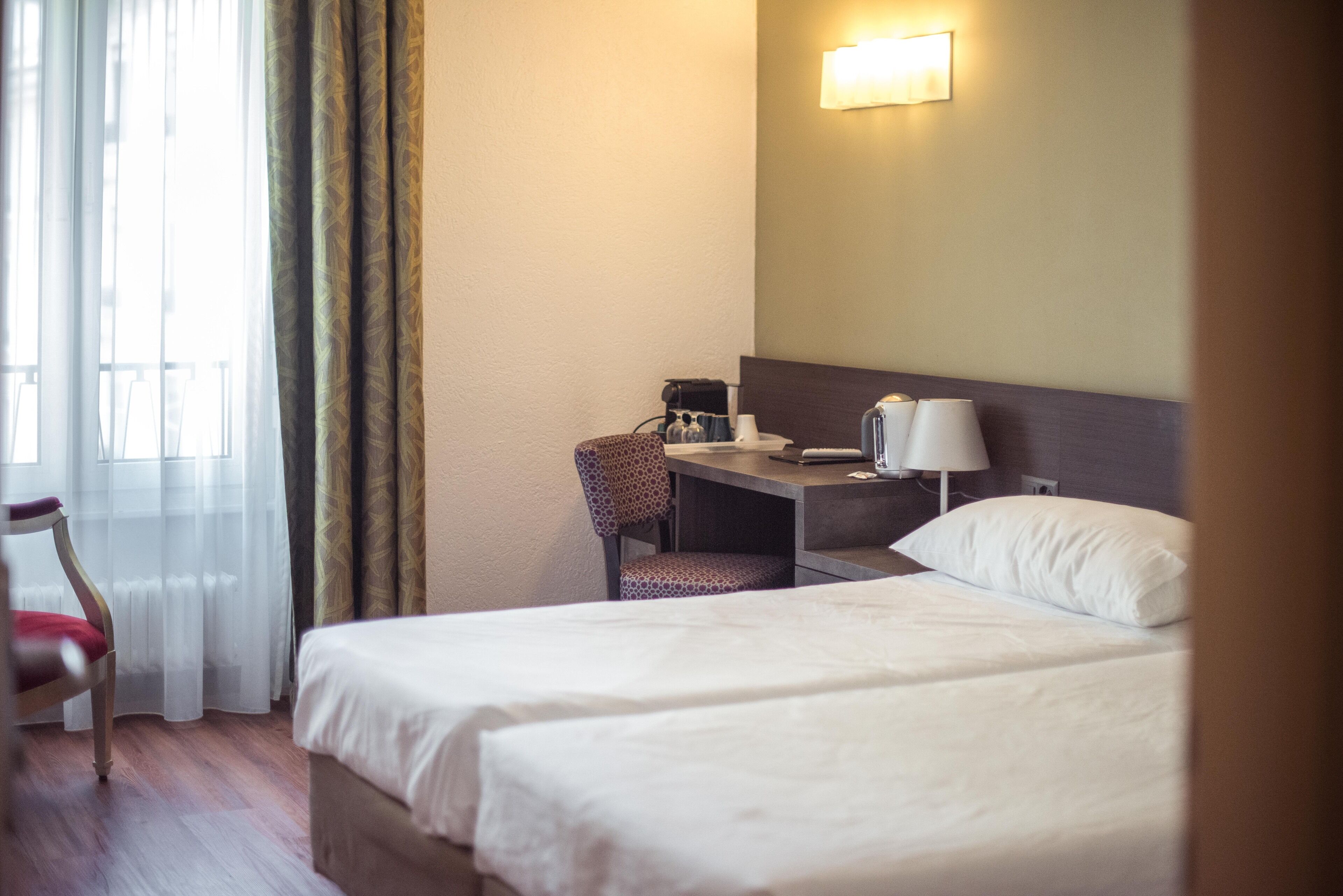 Une chambre d’hôtel avec deux lits, un bureau avec une lampe, une chaise et une fenêtre avec des rideaux.